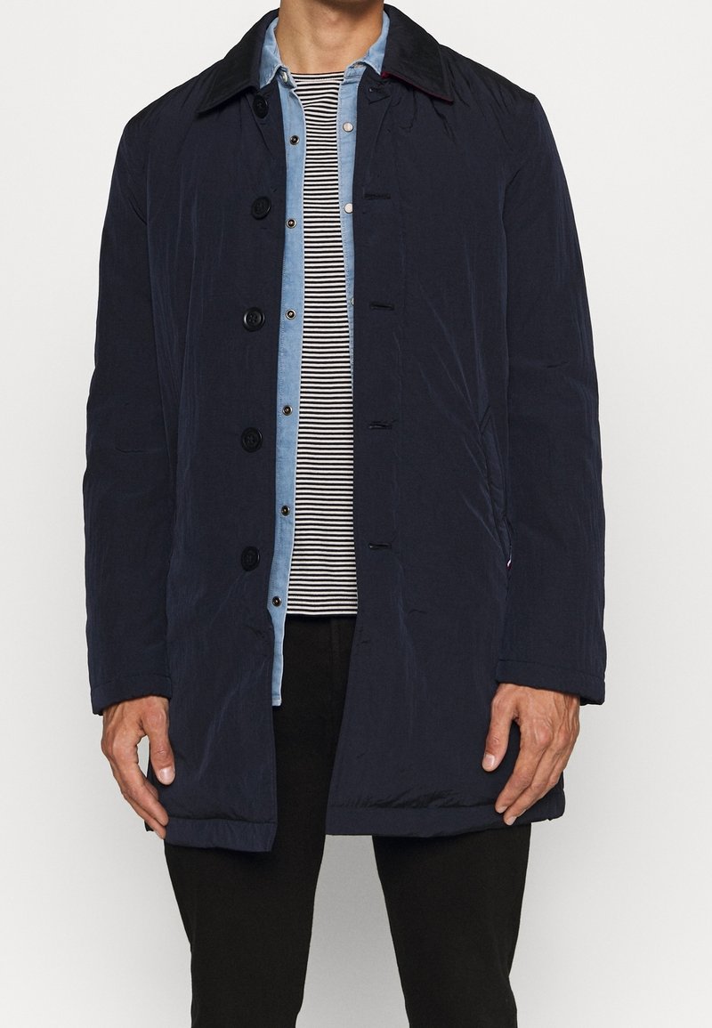 Marineblauer gesteppter Mantel mit Knopfverschluss, kontrastierendem Kragen und Seitentaschen, kombiniert mit einem hellblauen gestreiften Jeanshemd.