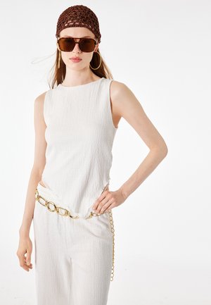 Mujer con top blanco sin mangas texturizado y pantalones, gafas de sol carey, pendientes de aro dorados, gorro marrón de punto y cinturón dorado de cadena.