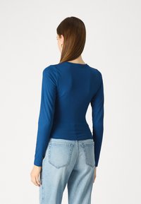 Μακρυμάνικο μπλουζάκι σε σκούρο μπλε, εφαρμοστή σχεδίαση με στρογγυλή λαιμόκοψη. Φτιαγμένο από απαλό, ελαστικό ύφασμα. Συνδυάζεται με ανοιχτό μπλε ψηλόμεσο τζιν.