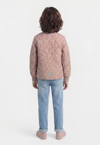 Kind mit lockigem Haar, das steht, trägt eine pinkfarbene gesteppte Jacke, hellblaue hochgekrempelte Jeans und rosa Schuhe, ist von hinten auf einem weißen Hintergrund zu sehen.