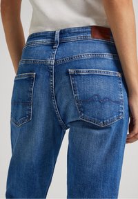 Blå denimjeans med hög midja, framträdande bakfickor med sömdetaljer och en brun läderetikett på midjebandet.