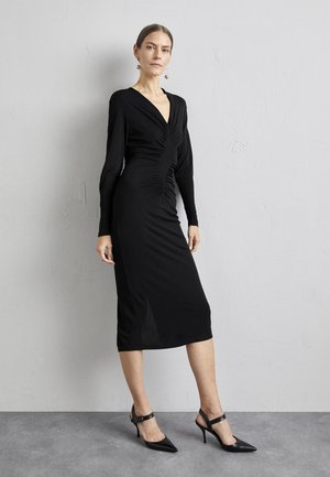 Μαύρο μακρυμάνικο φόρεμα με V-λαιμόκοψη, ραμμένο κορσέ και μήκος ως το γόνατο. Φοριέται με μαύρες μυτερές γόβες, που διαθέτουν λεπτό λουράκι στον αστράγαλο.