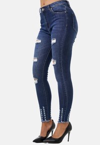 Jeans de mezclilla oscuros y ajustados, con desgastes y roturas, dobladillos deshilachados y costuras decorativas a lo largo de los lados, combinados con unos tacones altos negros.