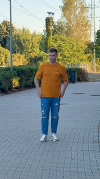 Pomarańczowa koszulka z krótkim rękawem, niebieskie zniszczone dżinsy oraz białe trampki z szarymi akcentami. W tle nawierzchnia i zieleń.