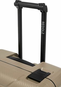 Poignée noire extensible avec prise texturée attachée à une valise rigide beige avec poignée de transport noire intégrée et verrou à loquet.