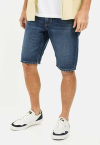 Mørkeblå denimshorts i midt-lår længde med slidte kanter. Sammen med hvide sneakers med marineblå og guld detaljer.