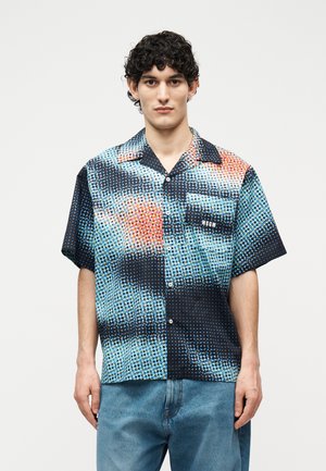 Young man with short curly hair wears a short-sleeve button-up shirt with blue, black, and orange dot pattern and light blue jeans.