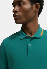 Polo shirt teal con colletto a pieghe, due bottoni e accentuazione arancione sul colletto. Texture liscia e design aderente.