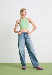 Jovem com cabelo loiro curto, vestindo um top verde curto, jeans azuis largos e ténis roxos, em pé sobre um piso verde contra uma parede branca.