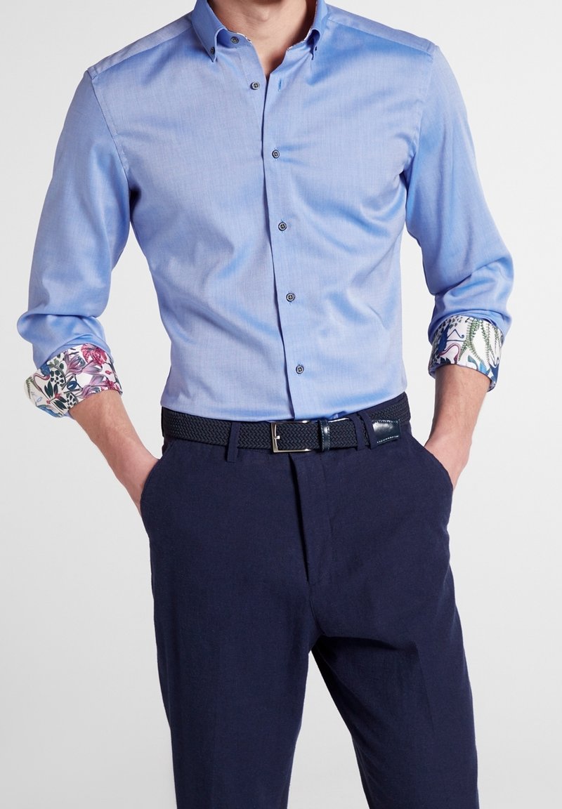 Homme portant une chemise bleu clair boutonnée avec motif floral aux manches retroussées et un pantalon bleu marine foncé avec ceinture noire, mains dans les poches.