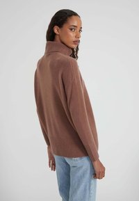 Frau mit lockigem Haar, die einen braunen Rollkragenpullover und hellblaue Jeans trägt, seitlich vor einem schlichten grauen Hintergrund steht.