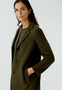 Manteau en mélange de laine vert olive, avec un col à revers, fermeture à trois boutons et poches latérales. Texture douce avec une coupe ajustée légèrement cintrée.