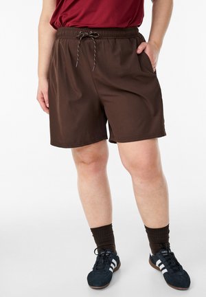 Osoba w brązowych spodenkach na sznurek, ciemnych skarpetkach i czarnych trampkach z białymi paskami, stojąca z jedną ręką w kieszeni.