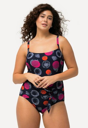Kvinde med krøllet hår iført en navy badedragt med rødt og pink frugtmønster, stående med hænderne foldet foran på en hvid baggrund.