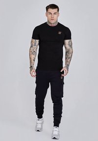 Zwart T-shirt met korte mouwen en een logo op de borst, gecombineerd met zwarte cargobroek. Het model draagt witte sportschoenen.