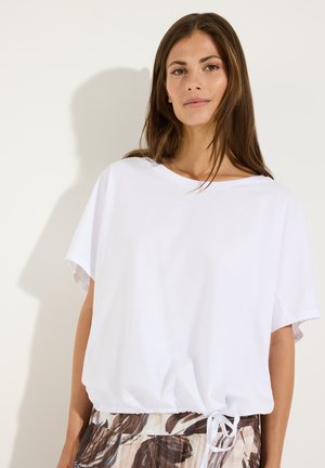 Frau mit langen braunen Haaren trägt ein lockeres weißes Kurzarmshirt mit Kordelzug am Saum und einen bedruckten Rock, steht vor einem einfarbigen hellen Hintergrund.