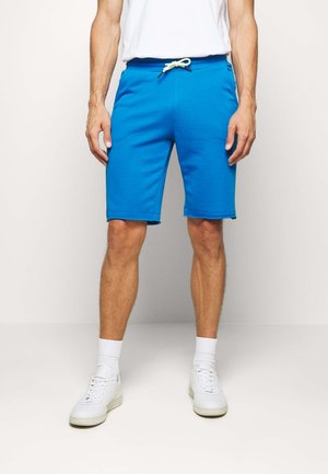 Homme portant un short bleu vif avec un cordon, des chaussettes blanches, des baskets blanches et un t-shirt blanc, debout devant un fond uni.