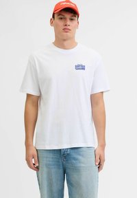 Jeune homme portant un t-shirt blanc avec le texte bleu "Handyman" sur la poitrine, un jean bleu clair et une casquette rouge avec un texte blanc.