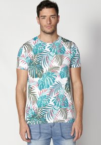 Camiseta de manga corta fabricada en algodón, con una base blanca y un vibrante estampado de hojas tropicales en tonos azul verdoso, verde y rosa.