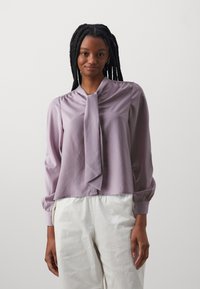 Lavender silk blouse with a tie collar, long sleeves with elastic cuffs, smooth texture, and a cropped design. Paired with white pants.