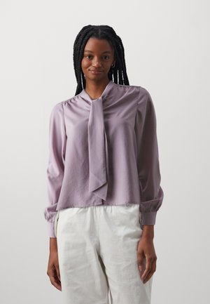 Lavender silk blouse with a tie collar, long sleeves with elastic cuffs, smooth texture, and a cropped design. Paired with white pants.