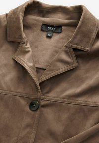 Veste en daim marron avec large col et un seul bouton noir, avec une étiquette de la marque "NEXT" visible et une étiquette "Made in China" à l'intérieur de l'encolure.