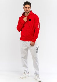 Rote Zip-Up-Hoodie mit einem großen schwarzen Kreis auf der Brust, kombiniert mit weißen Jogginghosen mit schwarzen Druckdetails. Weiße Turnschuhe runden den Look ab.