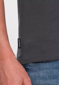 Tmavě šedé bavlněné tričko s krátkým rukávem, s černou etiketou na levém bočním švu. Detail ruky na denimu.