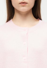 Γυναίκα που φοράει ένα απαλό ροζ ζακετάκι με κουμπιά, εμφανίζοντας το ντεκολτέ και τρία κουμπιά σε κοντινή λήψη.