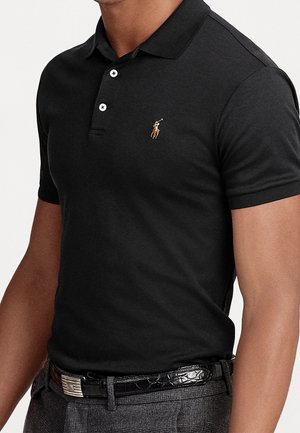 Mand iført tætsiddende sort poloshirt med lille broderet polospiller-logo, kombineret med grå ternede bukser og sort læderbælte.