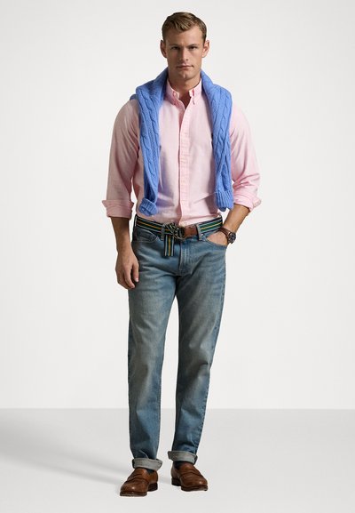 Sweater de malha azul claro sob uma camisa listrada rosa, combinado com calças jeans azul claro e sapatos de couro castanho. Apresenta um cinto listrado.