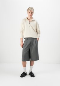 Beige vetoketjupaita logolla, harmaat laskostetut shortsit ja mustat loaferit, yhdistettynä valkoisiin miesten sukkiin. Yksinkertainen valkoinen tausta.