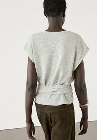 Light grey, short-sleeve knit top with a twisted back design. Paired with dark brown pants. Smooth texture, casual fit, minimalistic style.