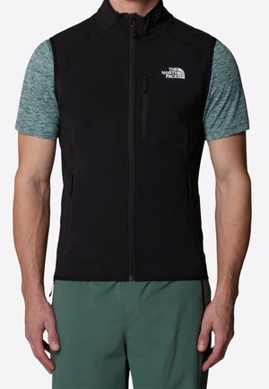 Mann trägt eine schwarze Zip-up-Weste über einem blauen kurzärmeligen Hemd und grünen Hosen, die Weste hat das The North Face-Logo auf der Brust.