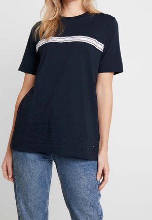 Kobieta w granatowym T-shircie z białym paskiem z logo Tommy Hilfiger przez klatkę piersiową i niebieskich dżinsach, stojąca na jednolitym tle.