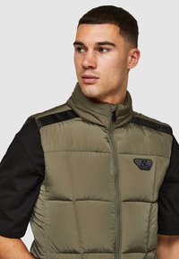 Olijfgroene puffervest met een hoge kraag, ritssluiting aan de voorkant en zwarte schouderaccenten. Voorzien van een geborduurde textuur en een merkembleem.