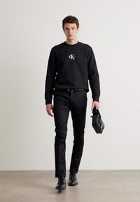 Sweatshirt noir avec logo, pantalons noirs et chaussures. Porte un sac rectangulaire noir. Modèle avec des cheveux courts et texturés. Fond neutre.