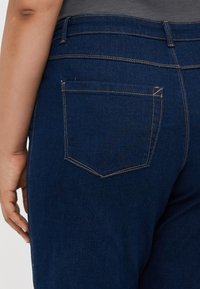 Mørkeblå denimjeans med en synlig baklomme. Teksturen er glatt med gule sømmer langs sømmene. Ingen tilleggsmaterialer.