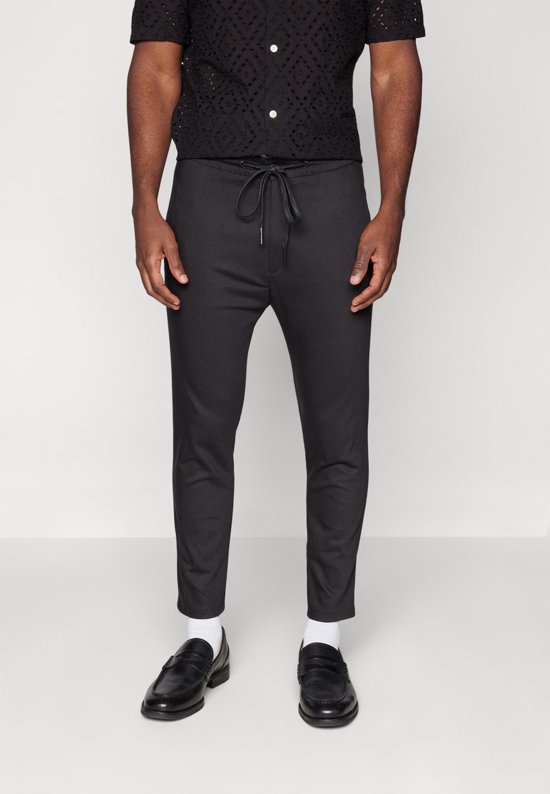 Pantalons de jogging noirs ajustés avec une taille élastique et un cordon de serrage. Texture lisse, jambes fuselées, associés à des mocassins noirs et des chaussettes blanches.