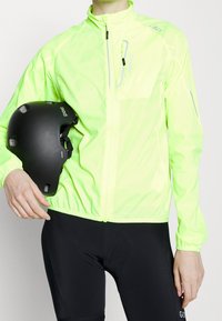Veste de cyclisme jaune fluorescent avec fermeture éclair frontale, accents réfléchissants, poignets élastiques et silhouette ajustée ; casque noir tenu à la main.