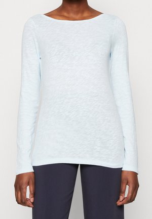 Frau in einem hellblauen Langarmshirt mit Bootsausschnitt und dunkler Hose, stehend vor einem einfarbigen Hintergrund.