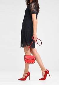 Červená kožená crossbody taška se zdobením, spárovaná s černými krajkovými šaty a červenými vysokými podpatky s páskem a špičatou špičkou.