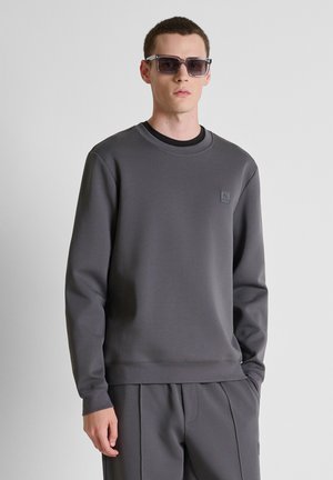 Jonge man die een grijze trui en bijpassende broek draagt, met een donkere zonnebril, staand tegen een effen lichte achtergrond.