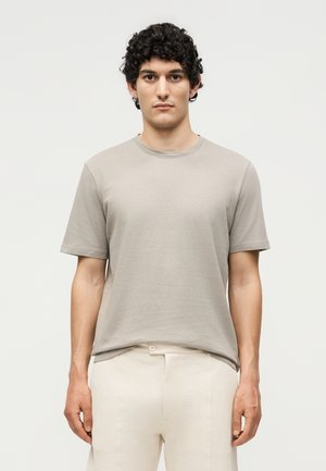 Νέος άνδρας με σγουρά σκούρα μαλλιά, που φοράει μπεζ κοντομάνικο πουκάμισο και εκρού παντελόνια, στέκεται μπροστά σε ένα απλό φωτεινό φόντο.