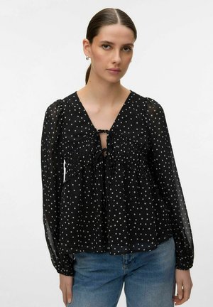 Femme aux cheveux bruns attachés, portant un chemisier noir transparent à pois avec des manches longues et un jean bleu, sur un fond blanc uni.