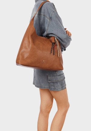 Donna con giacca di jeans e gonna che tiene una grande borsa a tracolla in pelle marrone con nappine decorative.
