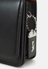 Černá kožená taška s hladkým povrchem, s kovovými prvky ve stříbrné barvě a detailem klíčenky. Šité okraje dodávají strukturní a rafinovaný vzhled.