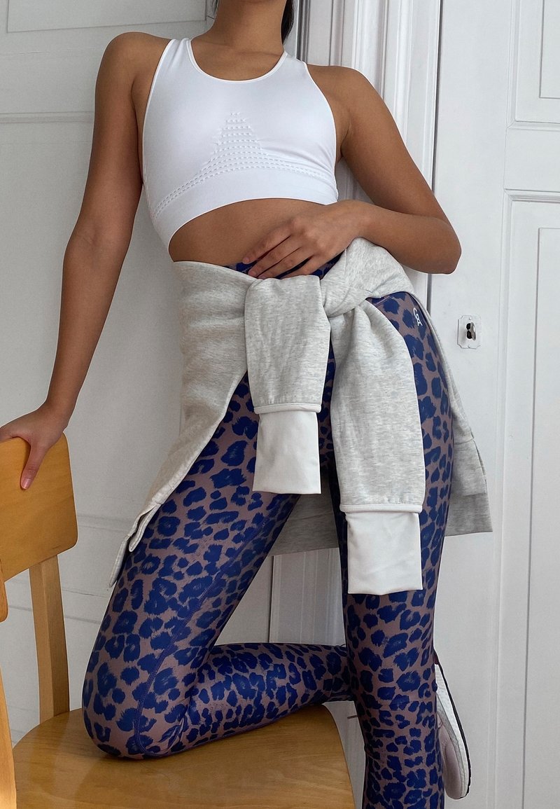 Soutien-gorge de sport blanc avec des détails texturés, sweat-shirt gris noué à la taille, et leggings léopard bleu avec des sections transparentes.