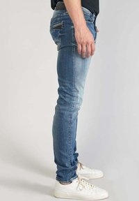 Jeans aderenti blu con finitura délavé, dotati di una tasca posteriore con cerniera, abbinati a sneaker bianche. Tessuto denim liscio.