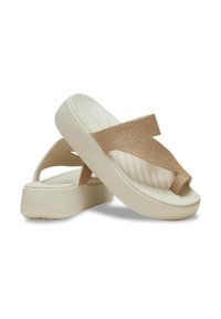 Beige platform flip-flops met een getextureerde gouden glitterschoenband. De voetbed bevat een ribbelontwerp voor grip en is aan de zijkant gemarkeerd.